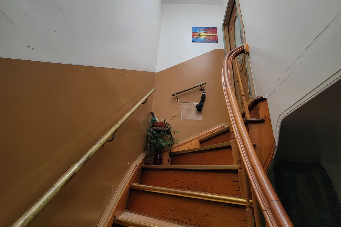 A stair hall before painting: white upper walls over a tan lower wall with a bare plaster patch, a brass handrail and a curved wooden banister.