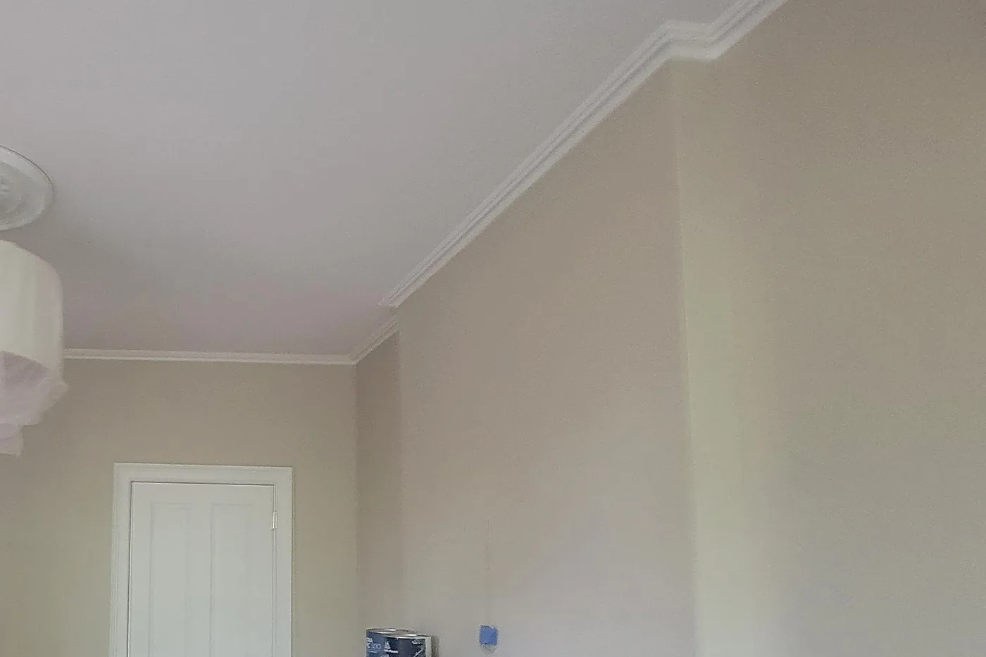 A parlor mid-job: warm greige walls meet a white crown molding and a white ceiling in straight, clean lines, the crown stepping out around the chimney breast, with blue tape and paint cans at the bottom of the frame.